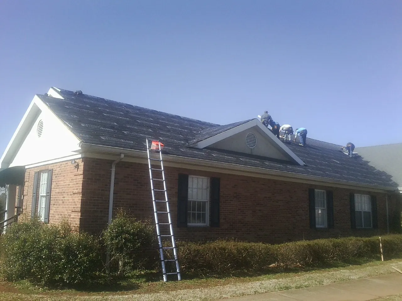 Crew tearing shingles off a brick ranch roof with a ladder set — Metal Roof Repair work by our crew in Hordville