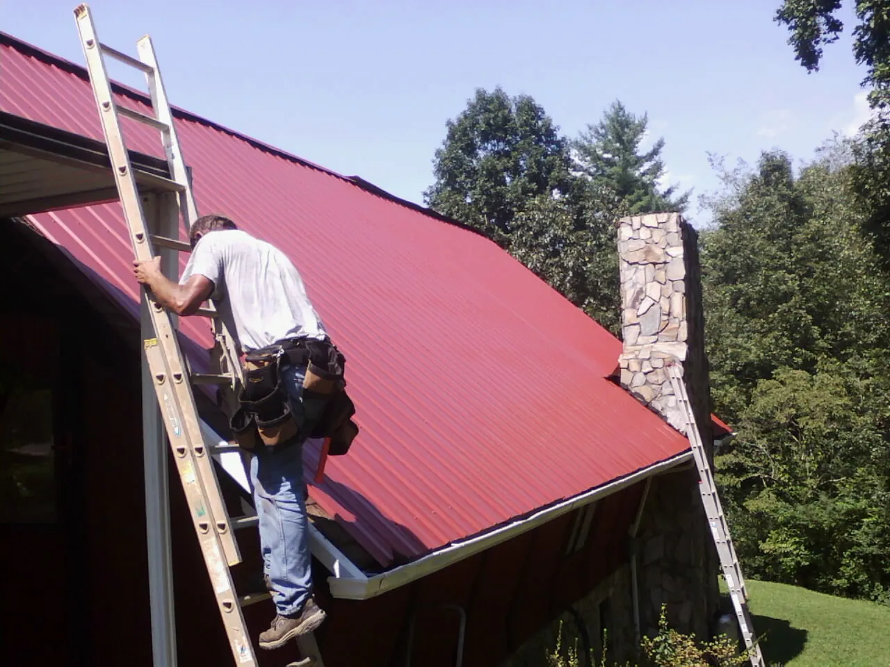 Roofer on a ladder beside a red metal roof and stone chimney — Soffit & Fascia Repair by Roofing Brothers in Hordville